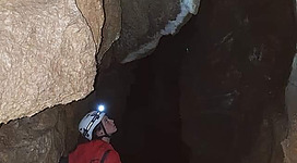 Eine Person in einem engen Spalt in einer Höhle steht. Sie trägt eine Kopflampe und ein rotes Outfit.
