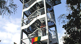 Aussichtswarte mit Heimkehrerkreuz in Waldhausen im Strudengau: Eine hohe, mehrstöckige Aussichtsplattform aus Metallgittern und Treppen, die inmitten von Bäumen und Grünflächen errichtet ist. Besucher können über die verschiedenen Etagen nach oben klettern, um von der Spitze aus einen Rundblick über die Umgebung zu genießen.