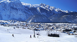 Eine schneebedeckte Landschaft mit Bergen im Hintergrund und einem kleinen Dorf. Skifahrer sind auf der Piste zu sehen, unter einem klaren blauen Himmel.