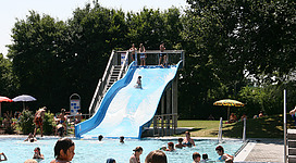 Freibad St.Florian in St. Florian: Ein Schwimmbad mit einer großen Rutsche, umgeben von Bäumen und Grünflächen. Zahlreiche Besucher, hauptsächlich Kinder und Jugendliche, sind im Wasser und auf der Rutsche zu sehen.