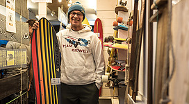 Ein Mann steht in einem Geschäft und hält ein buntes Surfbrett. Im Hintergrund sind verschiedene Skateboards und Sportartikel zu sehen.