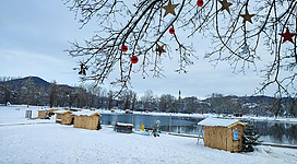 Ein winterlicher See mit schneebedecktem Ufer und kleinen Holzhütten. Am Baum hängen weihnachtliche Dekorationen in Rot und Gold.