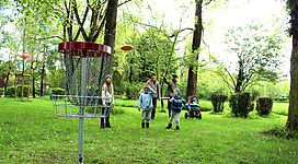 Discgolf in Braunau am Inn: Ein Frisbee-Korb in einem grünen Park mit Bäumen. Mehrere Personen, die offenbar Frisbee spielen, sind im Vordergrund zu sehen.