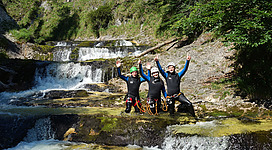 Canyoning bei den Gimbach Kaskaden in Steinbach am Attersee: Das Bild zeigt eine Gruppe von drei Menschen, die voller Begeisterung und mit erhobenen Armen in einem Fluss stehen, der von einem Wasserfall gespeist wird. Die Natur mit ihren grünen Bäumen und Felsen bildet eine malerische Kulisse für dieses Abenteuer.
