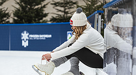 Eine Person schnürt ihre Schlittschuhe auf einer Eisbahn. Im Hintergrund sind Nadelbäume und eine blaue Wand zu sehen.