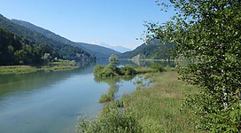 Ein ruhiger Fluss umgeben von grünen Wiesen und Bergen. Der Himmel ist klar und die Landschaft ist friedlich.