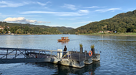 Donaufähre Grein in Grein: Eine ruhige Umgebung am Ufer eines Sees oder Flusses. Ein kleines Motorboot mit einer Holzplattform fährt über das ruhige Gewässer. Am Ufer befindet sich ein Bootsanleger mit Sitzmöglichkeiten. Die Landschaft ist von Wäldern und Bergen geprägt.