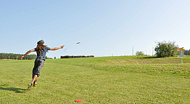 Discgolf Karlingberger Gut in Perg: Eine Person wirft ein rotes Frisbee über eine große grüne Rasenfläche. Sie trägt dunkle Kleidung und scheint sich in einer ländlichen Umgebung zu befinden, mit Bäumen und Sträuchern am Rand des Feldes.