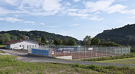 Tennisplatz in Wernstein am Inn: Eine große Lagerhalle aus Holz und Metall, umgeben von einer Metallzaun-Umrandung, befindet sich auf einer flachen Rasenfläche. Im Hintergrund sind bewaldete Berge und ein blauer Himmel mit weißen Wolken zu sehen.