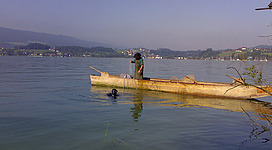 Fischer auf einem alten länglichen Holzboot im See, Taucher davor, im Hintergrund die Landschaft