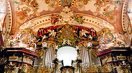 Zisterzienserstift und Kirche Wilhering in Wilhering: Ein reich verzierter Kirchenraum, dominiert von einem prachtvollen Orgelgehäuse. Die Wände sind mit detaillierten Fresken und Ornamenten geschmückt, die eine opulente Atmosphäre schaffen. Auf dem Altaraufsatz befinden sich weitere kunstvoll gestaltete Elemente. Das Gesamtbild vermittelt einen Eindruck von Schönheit, Pracht und Spiritualität.