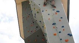 Ein Kletterer erklimmt eine hoch aufragende Kletterwand. Der Himmel ist teilweise bewölkt und die Wand ist mit bunten Griffen ausgestattet.