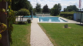 Freibad Wolfsegg in Wolfsegg am Hausruck: Dies zeigt eine ansprechende Außenanlage mit einem großen Schwimmbecken. Es gibt einen Gehweg, der durch einen gepflegten Garten mit Bäumen und Sträuchern führt. Der Boden ist mit Pflastersteinen ausgelegt. Im Hintergrund sind weitere Gebäude und Vegetation zu sehen, die eine angenehme, grüne Umgebung schaffen.