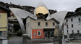 Ein modernes, skulpturales Tor aus Metall mit einem goldenen Element in der Mitte. Im Hintergrund sind Gebäude und eine bewölkte Himmel zu sehen.