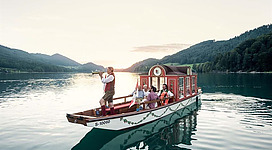 Ein traditionelles Boot fährt auf einem ruhigen See, umgeben von Bergen. Die Passagiere genießen die Aussicht, während der Kapitän musiziert.