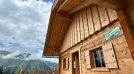 Eine gemütliche Holzhütte mit einer einladenden Terrasse. Im Hintergrund sind Berge und ein blauer Himmel zu sehen.