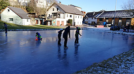Eislaufplatz in Lembach im Mühlkreis: Eine Gruppe von Menschen, die bei kaltem Wetter auf einer gefrorenen Wasserfläche Schlittschuh laufen. Sie tragen Mäntel und Handschuhe und scheinen die Kälte und den Spaß auf dem Eis zu genießen. Im Hintergrund sind kleine Wohnhäuser und Gebäude zu sehen, die eine gemütliche, winterliche Atmosphäre vermitteln.