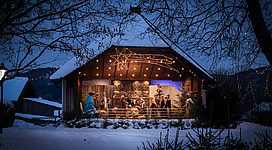 Auf dem Foto sieht man eine weihnachtliche Krippe mit überlebensgroßen Krippenfiguren. Die Krippe ist im Musikpavillon ausgestellt und es befinden sich am Foto auch Schnee.