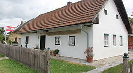Heimathaus Naarn im Machlande in Naarn im Machlande: Ein traditionelles Landhaus mit roten Ziegeldächern, weißen Wänden und grünen Fensterrahmen. Es befindet sich in einer ländlichen Umgebung mit einem Holzzaun und blühenden Pflanzen im Vordergrund. Das Gebäude wirkt einladend und gemütlich.