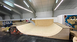Skatehalle in Ried im Innkreis: Eine große Halfpipe-Konstruktion in einem Innenraum mit beleuchteten Decken und Wänden. Es gibt Graffiti-Elemente an den Wänden, die darauf hinweisen, dass es sich um einen Raum für Aktivitäten wie Skateboarden handelt.