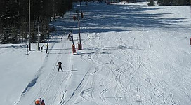 Eine schneebedeckte Piste mit Skifahrern und Lifts. Im Hintergrund sind Bäume und ein klarer blauer Himmel zu sehen.