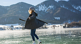 Eine Frau in einem schwarzen Mantel und Schlittschuhen gleitet fröhlich über eine gefrorene Seenfläche. Im Hintergrund sind schneebedeckte Berge und ein klarer blauer Himmel zu sehen.