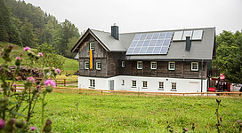 Ein modernes Haus mit Holzverkleidung und Solarpanelen steht in einer grünen Landschaft. Umgeben von Bäumen und Wiese, strahlt es ländlichen Charme aus.