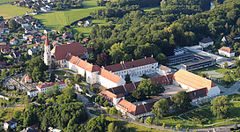 Schloss / Stift Ranshofen in Braunau am Inn: Ein historisches Gebäude mit traditioneller Architektur thront inmitten einer hügeligen, grünen Landschaft. Mehrere Türme und Dächer sind zu sehen, die auf ein großes Areal hinweisen. Umliegende Wohnhäuser und Wälder umgeben das Hauptgebäude, das den Mittelpunkt der Ansicht bildet.