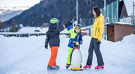 Eine fröhliche Familie auf einer Eislaufbahn im Winter. Zwei Kinder spielen mit einem Pinguin-Laufschlitten, während die Mutter neben ihnen steht.