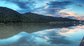 Donaukraftwerk Aschach in Aschach an der Donau: Eine friedliche Szenerie an einem ruhigen See, umgeben von bewaldeten Hügeln und einem malerischen Abendhimmel. Die Wasserfläche spiegelt die Landschaft und den Himmel in beruhigender Weise wider. Am Ufer sind einige Gebäude zu erkennen, die den See säumen.