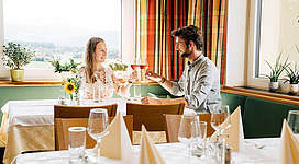 Ein Paar sitzt in einem gemütlichen Restaurant und stößt mit Gläsern an. Die Tische sind elegant gedeckt und es gibt Sonnenblumen auf dem Tisch.