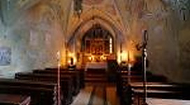 Eine ruhige Kapelle mit hölzernen Bänken und sanftem Kerzenlicht. Die Wände sind mit bunten, historischen Fresken geschmückt.