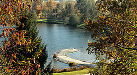 Ein ruhiger See umgeben von buntem Herbstlaub. Eine kleine Landzunge ragt ins Wasser hinaus.