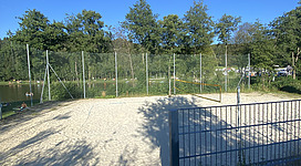 Beach-Volleyballplatz beim Badesee in Pramet: Ein Gitter aus Metallstangen umgibt eine grüne Rasenfläche. Dahinter befindet sich eine Hauswand mit Fensterläden. Hohe Bäume rahmen die Szene und ein strahlend blauer Himmel erstreckt sich darüber.