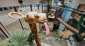 Eine große Giraffenskulptur aus Holz mit einer langen Zunge steht in einem modernen, mehrstöckigen Raum. Im Hintergrund sind Spielstrukturen und Pflanzen zu sehen.