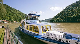 Donau-Schiff MS Lilofee in Obermühl an der Donau: Ein Personenfähre schwimmt auf einem ruhigen Gewässer, umgeben von bewaldeten Hängen. Die Fähre ist hell lackiert und hat eine Überdachung. Passagiere können an Bord gehen und von einer Seite des Gewässers zur anderen übersetzen.