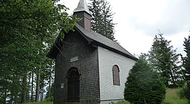 Richtberg-Kapelle in Weyregg am Attersee: Das Bild zeigt eine idyllische Holzkapelle, die von dichtem Wald umgeben ist. Das Gebäude hat ein markantes Dach mit einem Kreuz an der Spitze und befindet sich auf einer leicht erhöhten Position inmitten einer grünen Landschaft.