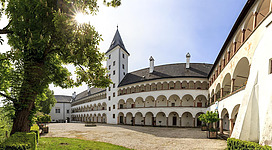 Ein historisches Gebäude mit weißer Fassade und einem hohen Turm. Der Innenhof ist von Arkaden umgeben und von Bäumen grünes umgeben.