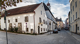 Heimathaus Vöcklabruck in Vöcklabruck: Ein klassisches Gebäude mit traditioneller Architektur, umgeben von herbstlichen Bäumen. Auf der Straße sieht man Kopfsteinpflaster und einige Autos. Der Himmel ist bewölkt, aber es scheint ein sonniger Tag zu sein.