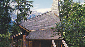 Eine charmante Holzkirche umgeben von dichten Bäumen und Bergen. Der hohe Spitzdach verleiht dem Gebäude einen markanten Look.