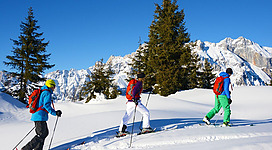 Drei Skifahrer wandern durch eine verschneite Landschaft. Im Hintergrund sind hohe Berge und ein klarer blauer Himmel zu sehen.