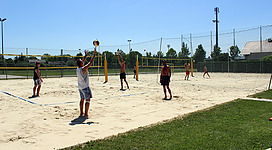 Volleyballplatz Braunau in Braunau am Inn: Auf dem Sportplatz spielen mehrere Personen Volleyball. Sie werfen den Ball über ein Netz, das zwischen zwei Pfosten gespannt ist. Der Boden besteht aus Sand, und die Umgebung ist von Bäumen und Gras umgeben.