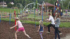 Motorikpark in Wernstein am Inn: Auf dem Bild ist ein abwechslungsreicher Spielplatz in einer ländlichen Umgebung zu sehen. Es gibt verschiedene Spielgeräte wie Schaukeln, Klettergerüste und bunte, abstrakte Kunstobjekte. Mehrere Kinder spielen und interagieren miteinander auf den Spielgeräten. Erwachsene Personen sind ebenfalls anwesend und beobachten das Geschehen. Die Umgebung ist grün und von Bäumen umgeben.