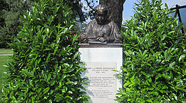 Marie von Ebner-Eschenbach - Denkmal in St. Gilgen: Das Bild zeigt eine Bronzebüste einer Frau in einem grünen, bewachsenen Umfeld. Die Büste steht auf einem Sockel, der mit einer Inschrift versehen ist. Die Umgebung ist von üppiger Vegetation geprägt, die die Büste einrahmt.