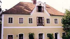 Schloss Mühlheim in Mühlheim am Inn: Ein charmantes Gebäude mit traditioneller Architektur, umgeben von Grünflächen und Topfpflanzen. Das zweistöckige Haus ist mit hellen Farben gestrichen und verfügt über ein markantes Dach mit einem Turm. An den Fenstern sind Fensterläden angebracht, und der Eingang ist mit Bögen verziert. Insgesamt vermittelt das Gebäude einen historischen und einladenden Eindruck.