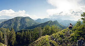 Ein Mensch sitzt auf einem Hügel und schaut auf eine malerische Berglandschaft. Die Umgebung ist grün mit Bäumen und hat einen klaren blauen Himmel.