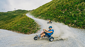 Eine Gruppe von Personen fährt mit Dreirädern eine kurvenreiche Strecke den Hügel hinunter. Staub wirbelt auf, während sie Spaß beim Fahren haben.