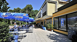 Ein Restaurant mit Tischen im Freien und blauen Sonnenschirmen. Im Hintergrund sind Bäume und ein Gebäude zu sehen.