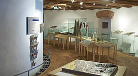 Turm 9 - Stadtmuseum Leonding in Leonding: Eine geräumige Ausstellungshalle mit Holzbalkendecke und mehreren Glasvitrinen, in denen verschiedene Objekte und Artefakte ausgestellt sind. Auf dem Boden steht eine große Landkarte oder geologische Karte, die das Interesse der Besucher wecken könnte. Die Beleuchtung ist warm und gemütlich, wodurch die Atmosphäre einladend wirkt.