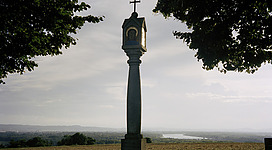 Fritz-Fröhlich-Wanderweg in Wilhering: Ein religiöses Symbol, bestehend aus einem Kreuz auf einer schlanken Säule, steht inmitten einer ländlichen Landschaft. Grüne Bäume rahmen die Szenerie ein, während sich im Hintergrund eine sanfte Hügellandschaft erstreckt.